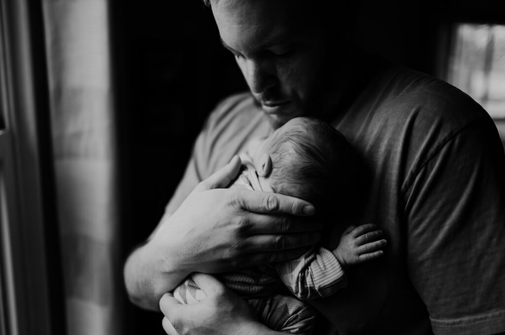 Father holding newborn in front of a window. 